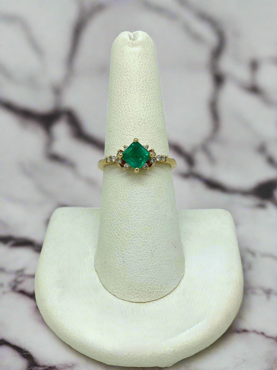 Gold ring with a princess cut Emerald and Diamond accents on a white stand